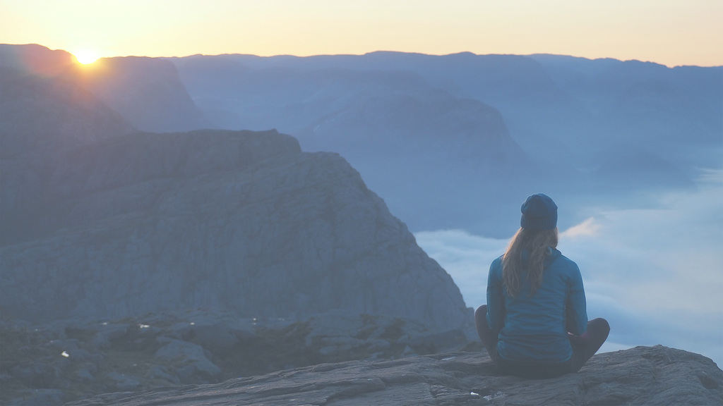Eine Person sitzt im Schneidersitz auf einem Felsen und blickt in die Weite eines Fjordes.